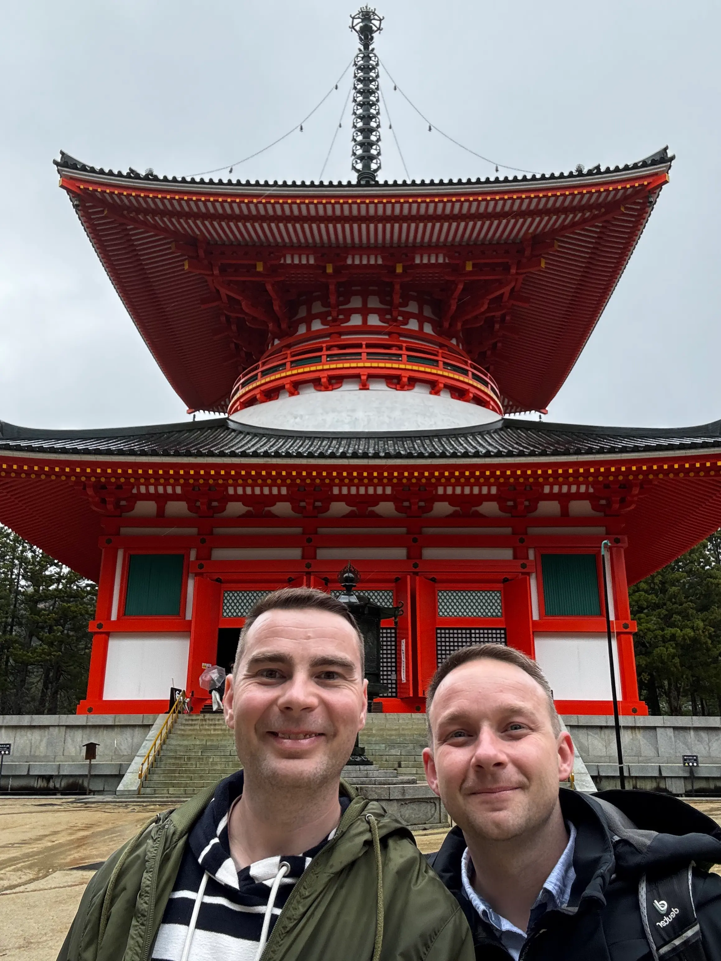 Před červenou japonskou svatyní Konpon Daito s velkou střechou a věží stojí dva muži, obloha je zatažená.