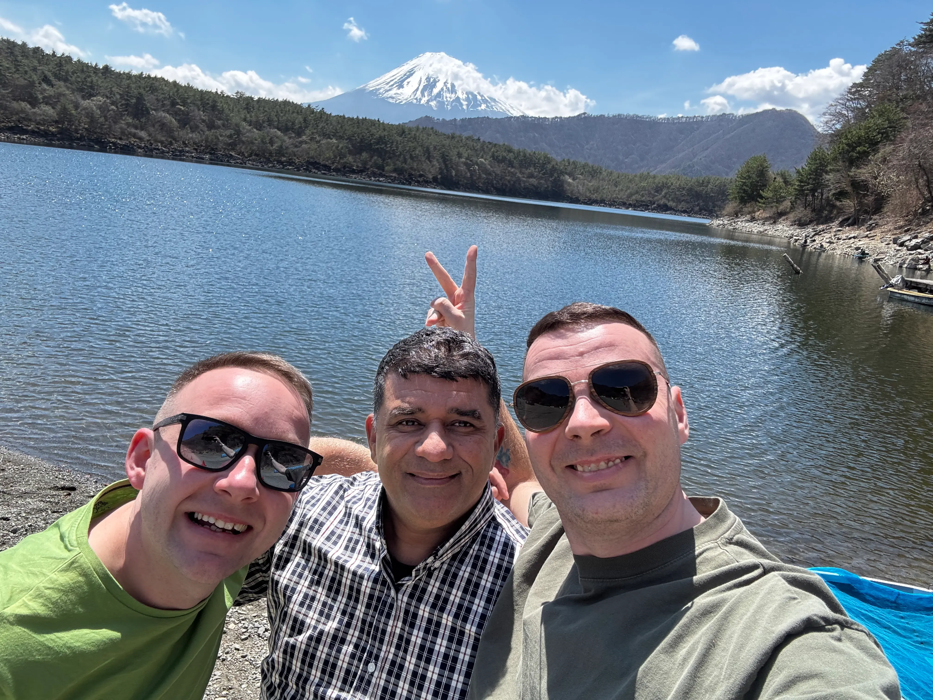 Tři muži na selfie u jezera Saiko, v pozadí zasněžená hora Fuji a lesnaté svahy. Jeden muž je řidič, dva další jsou turisté.