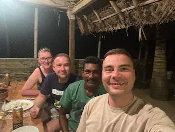 Skupina přátel sedí venku u stolu v zahradním altánu, selfie zblízka s úsměvy.