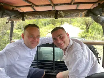 Dva muži v bílých košilích se usmívají pod přístřeškem u auta na safari na Srí Lance, venku v zeleni.