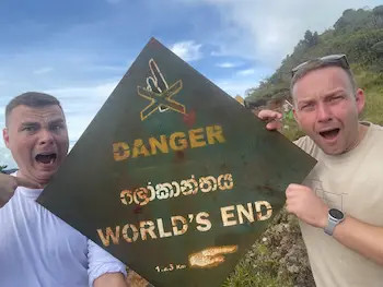 Dva muži drží ceduli s nápisem „DANGER“ a „WORLD’S END“, venku u lesa - Hortnovy planiny na Srí Lance.