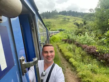 Muž v bílém tričku a s batohem pózuje na fotku ze dveří v jedoucím vlaku na Srí Lance, krajina s loukami a zelení.