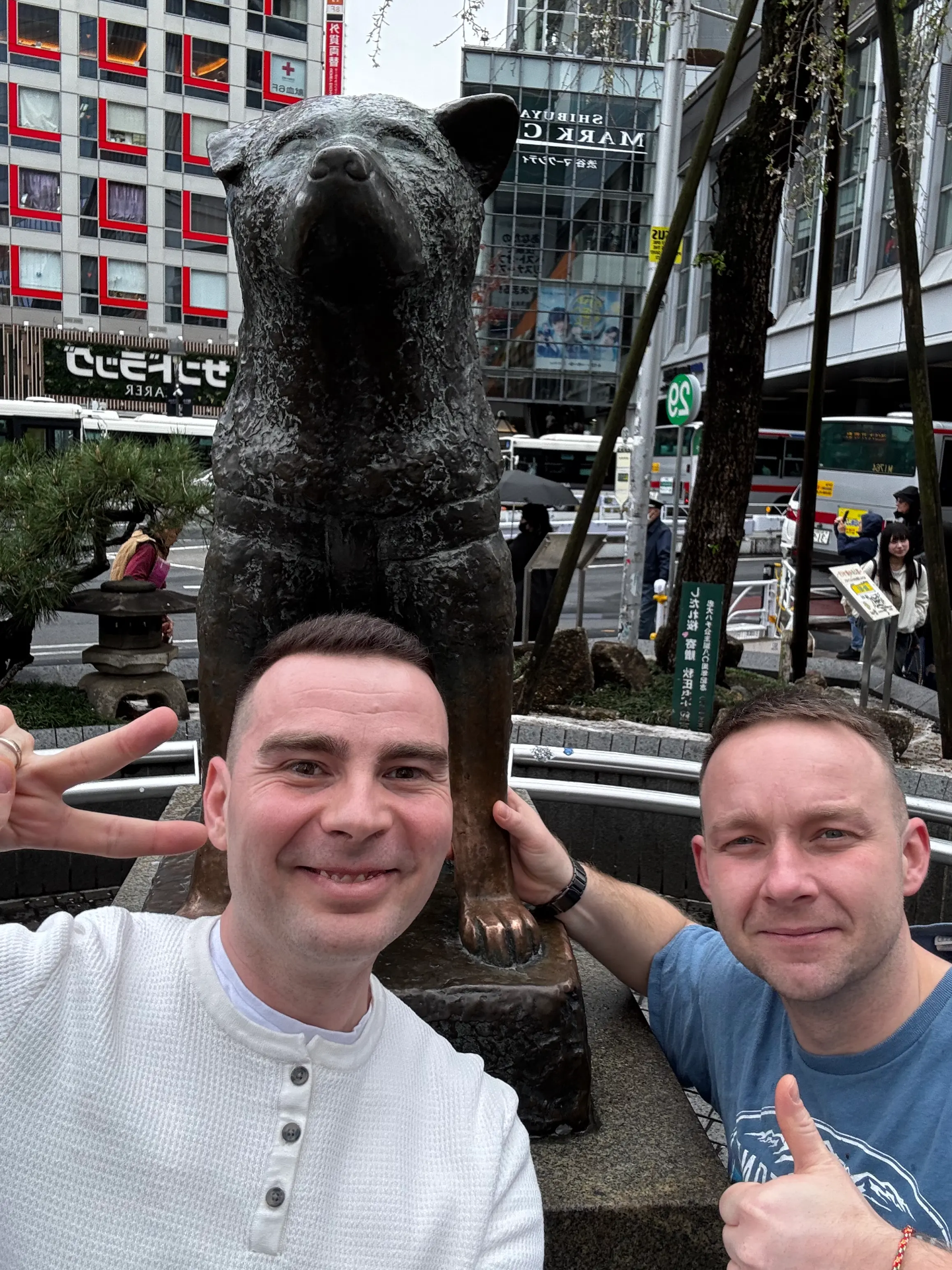 Dva muži pózují u bronzové sochy psa Hachiko, v pozadí městské budovy a auta.