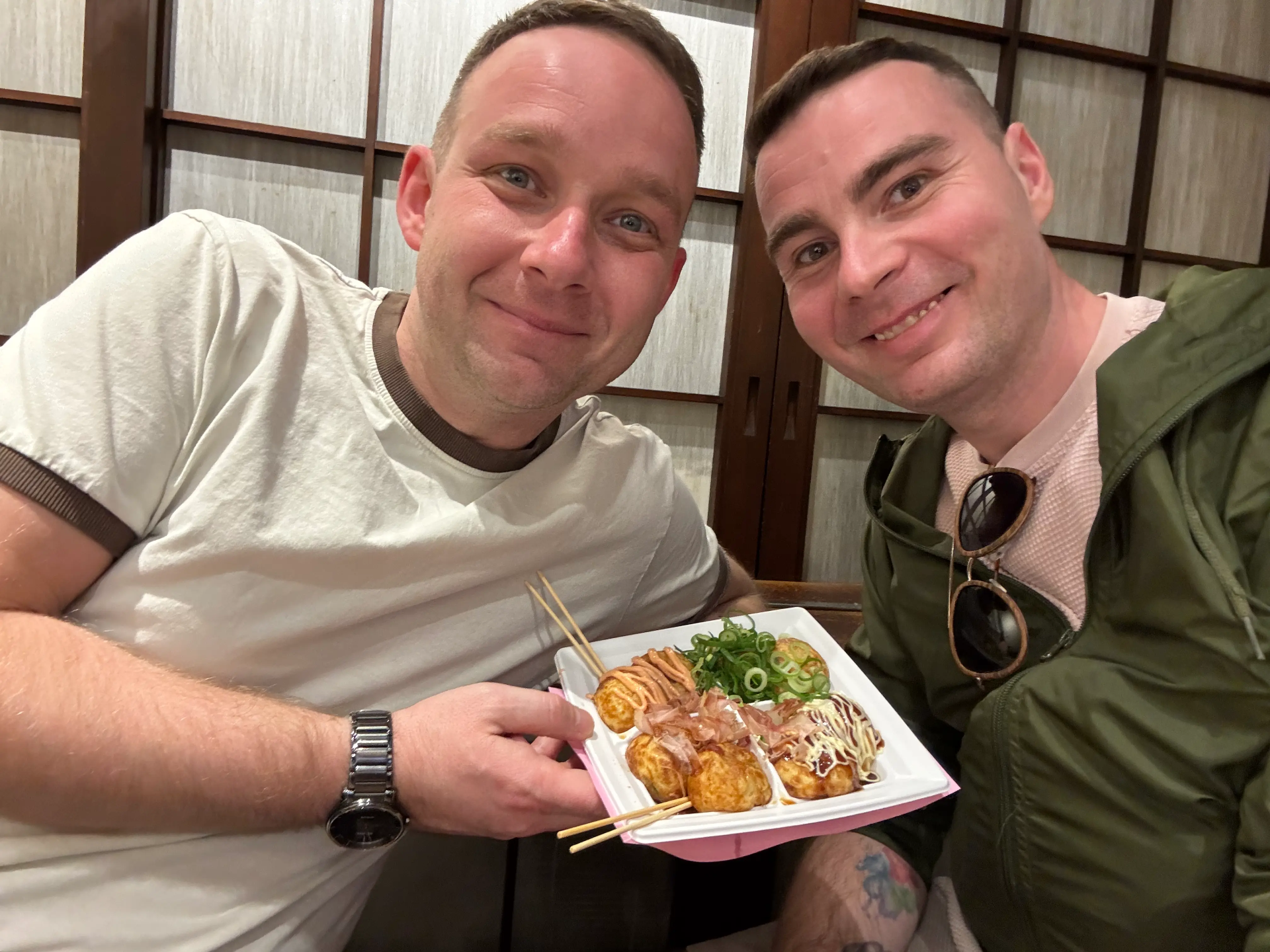 Dva muži se usmívají u stolu, drží krabičku s takoyaki a zelenou cibulkou.