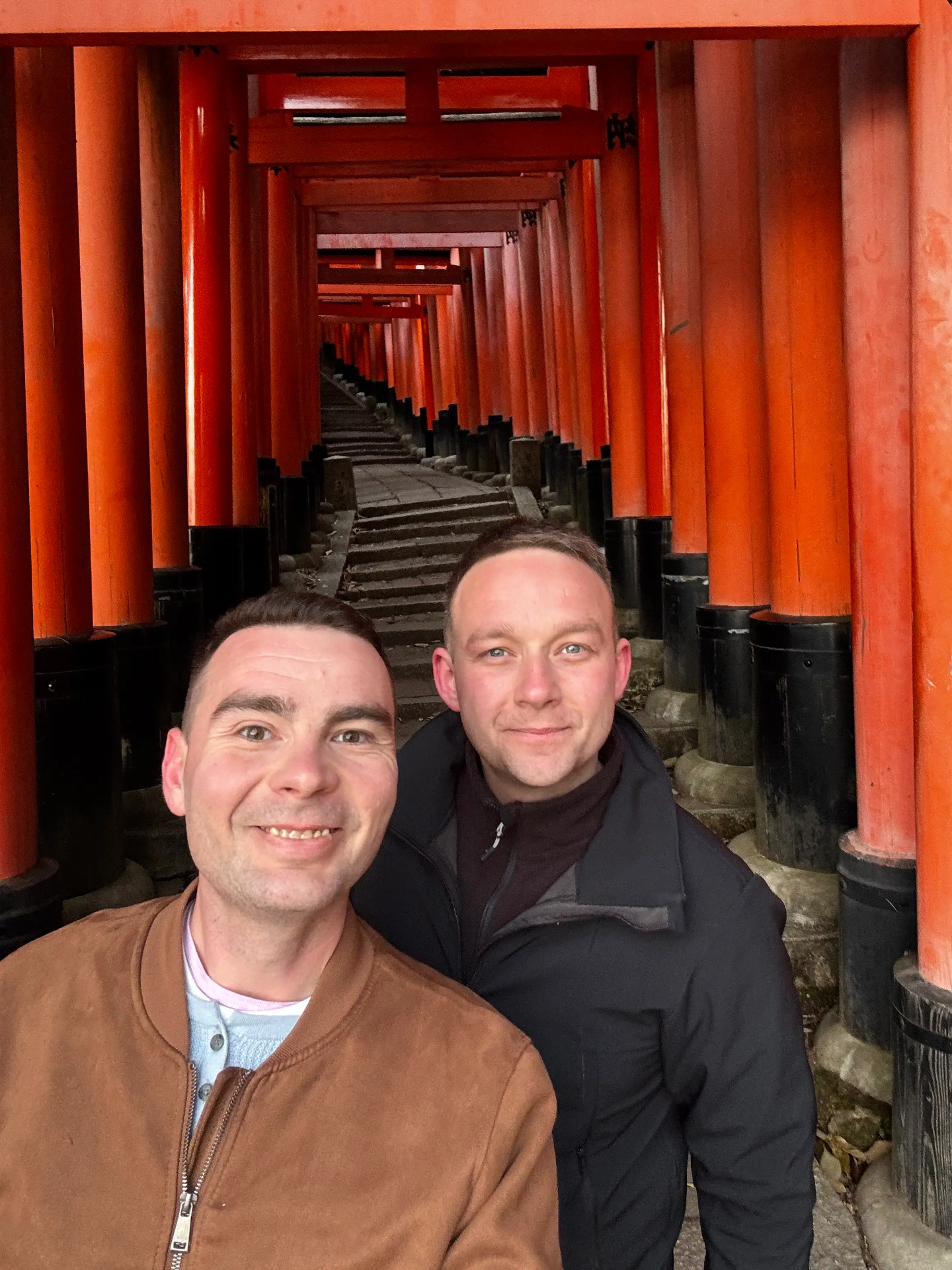 Dva muži v popředí pózují selfie v dlouhé chodbě s červenými torii a kamennými schody.