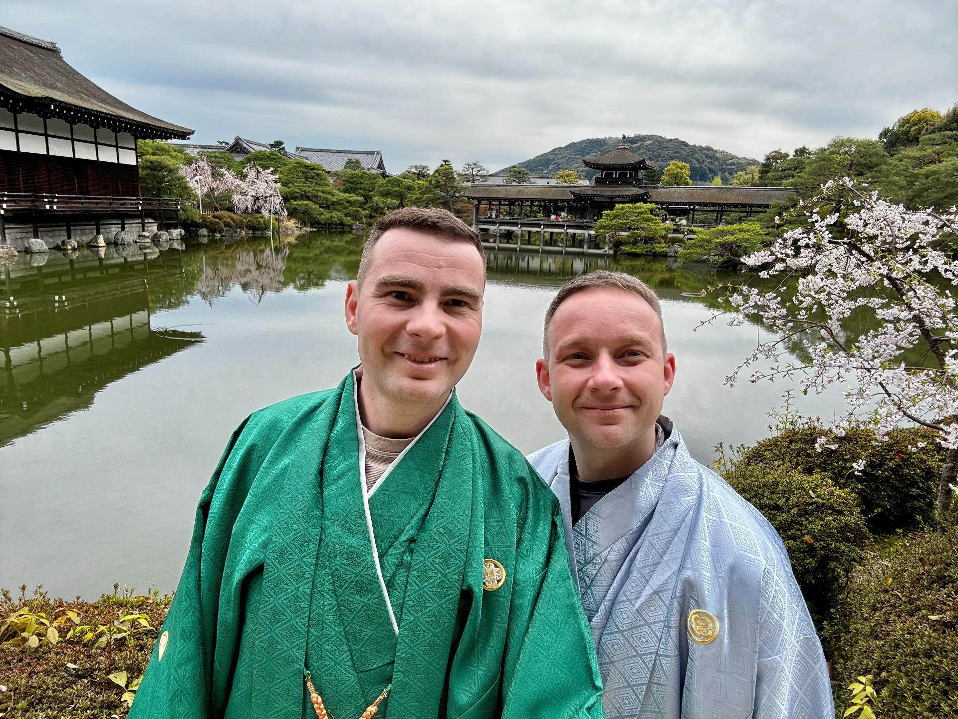 Dva muži v tradičních rouchách pózují u rybníka s japonským chrámem a třešňovými květy.