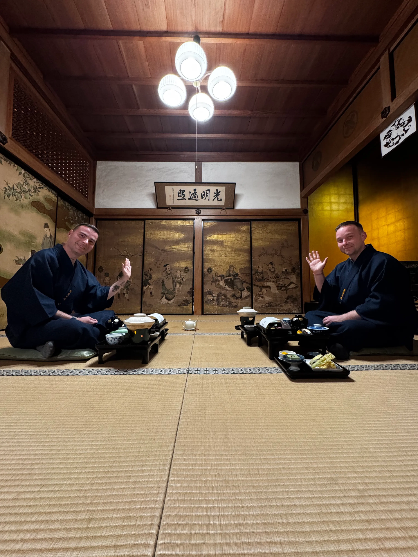 Dva muži v tradičních kimonech sedí na tatami, zdraví, před nimi čajové nádobí.