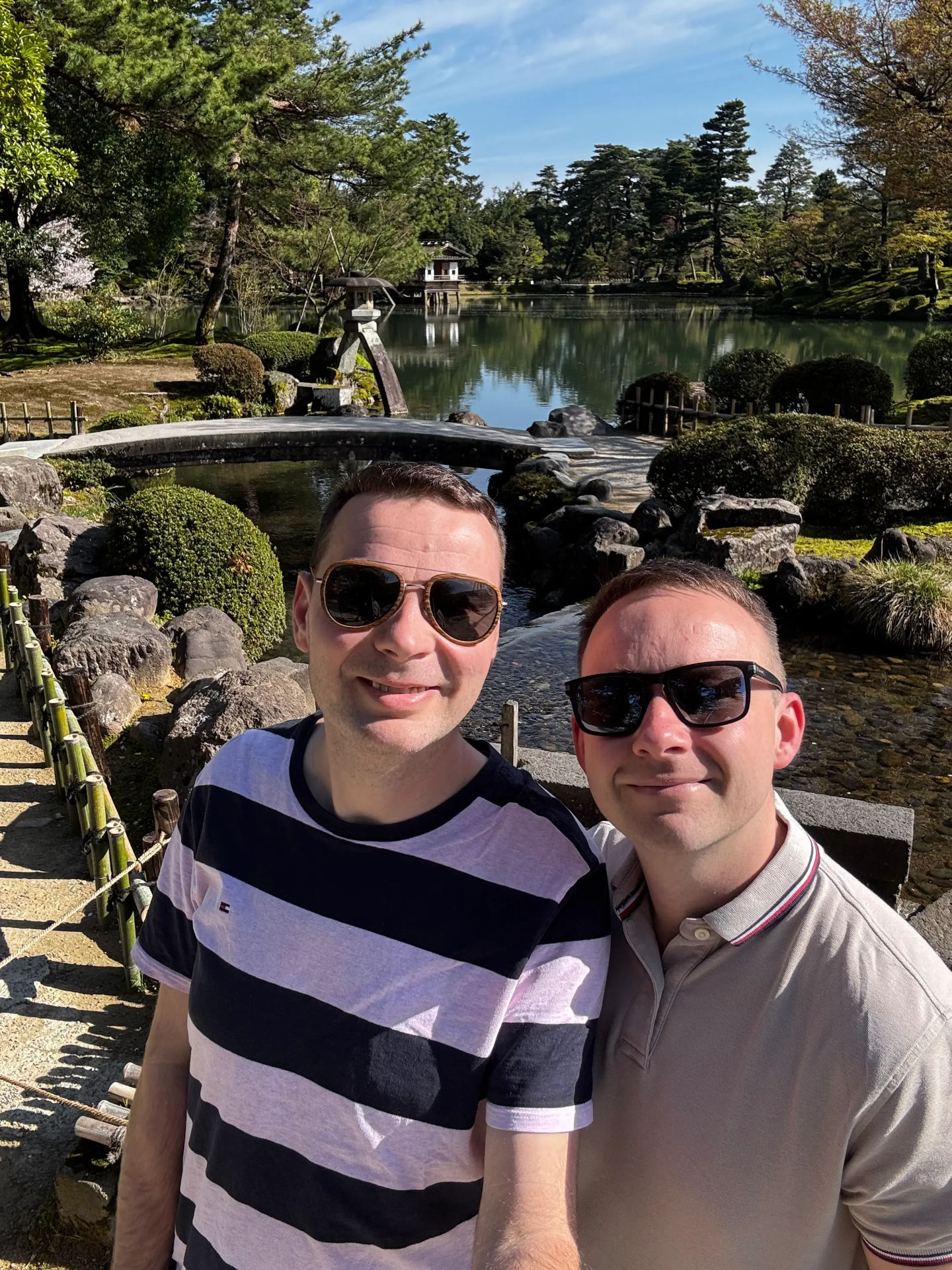 Dva muži v slunečních brýlích pózují u rybníka s kamennou fontánou a zelenými stromy.